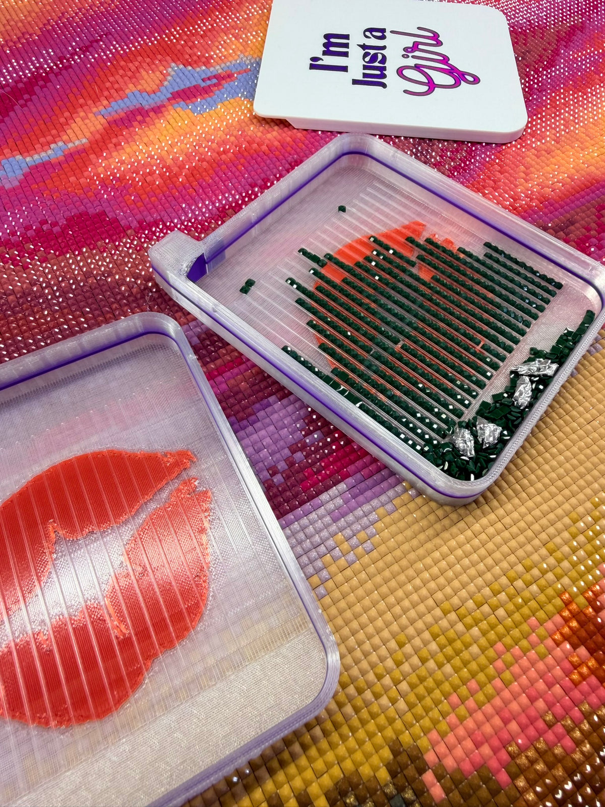 Close-up of two translucent lavender diamond painting trays resting on a colorful, partially completed diamond art canvas. One tray contains rows of dark green drills with a few orange and silver drills mixed in, while the other features a large orange-red lips design. A white rectangular accessory at the top reads ‘I’m Justa Girl’ in purple and pink lettering