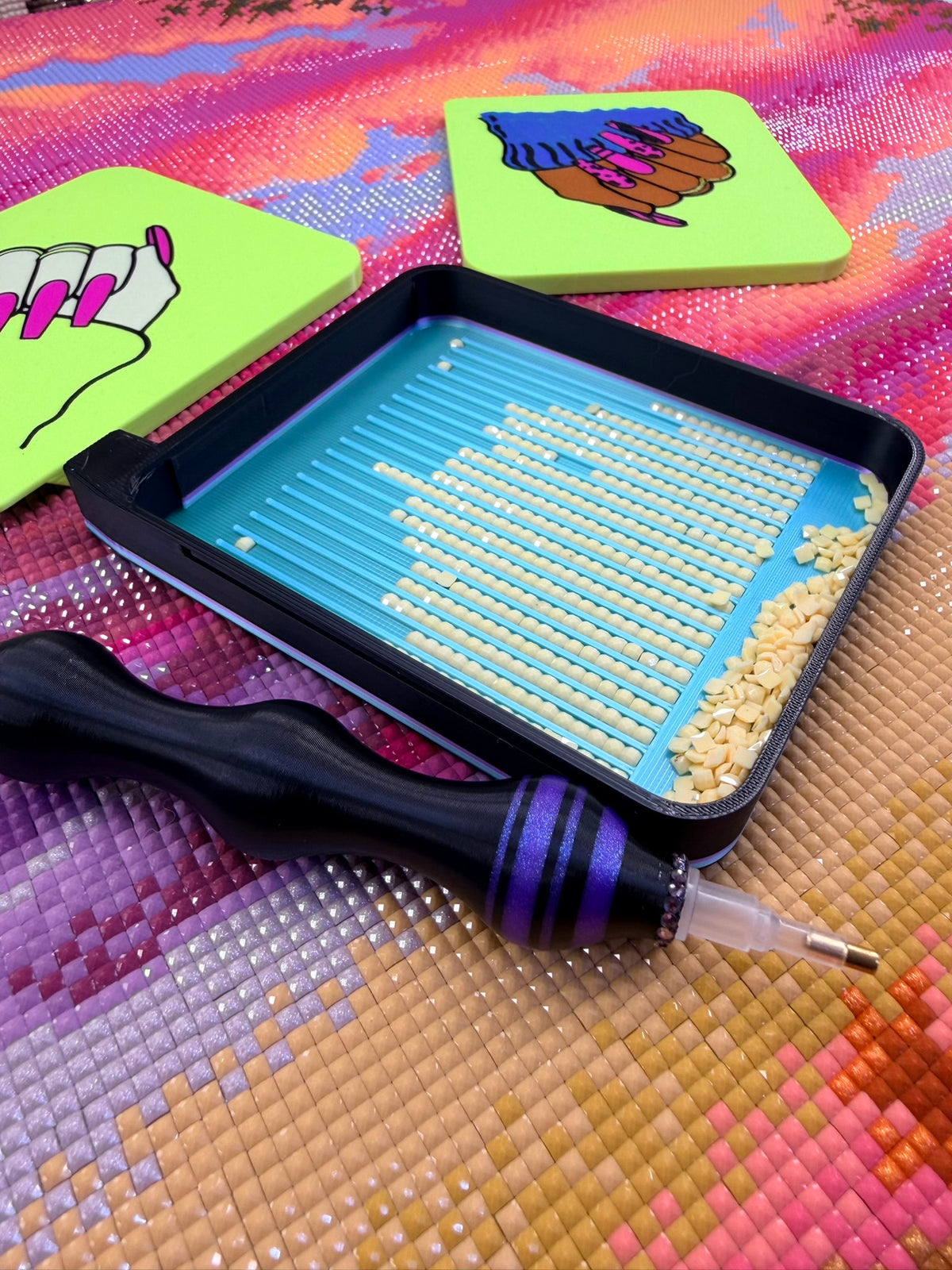 Close-up of a black and purple diamond painting tray filled with rows of pale yellow drills on a colorful diamond art canvas. A black ergonomic diamond painting pen with purple accents rests in front of the tray. Two bright lime green tray lids sit behind it, each decorated with illustrated hands featuring long pink nails