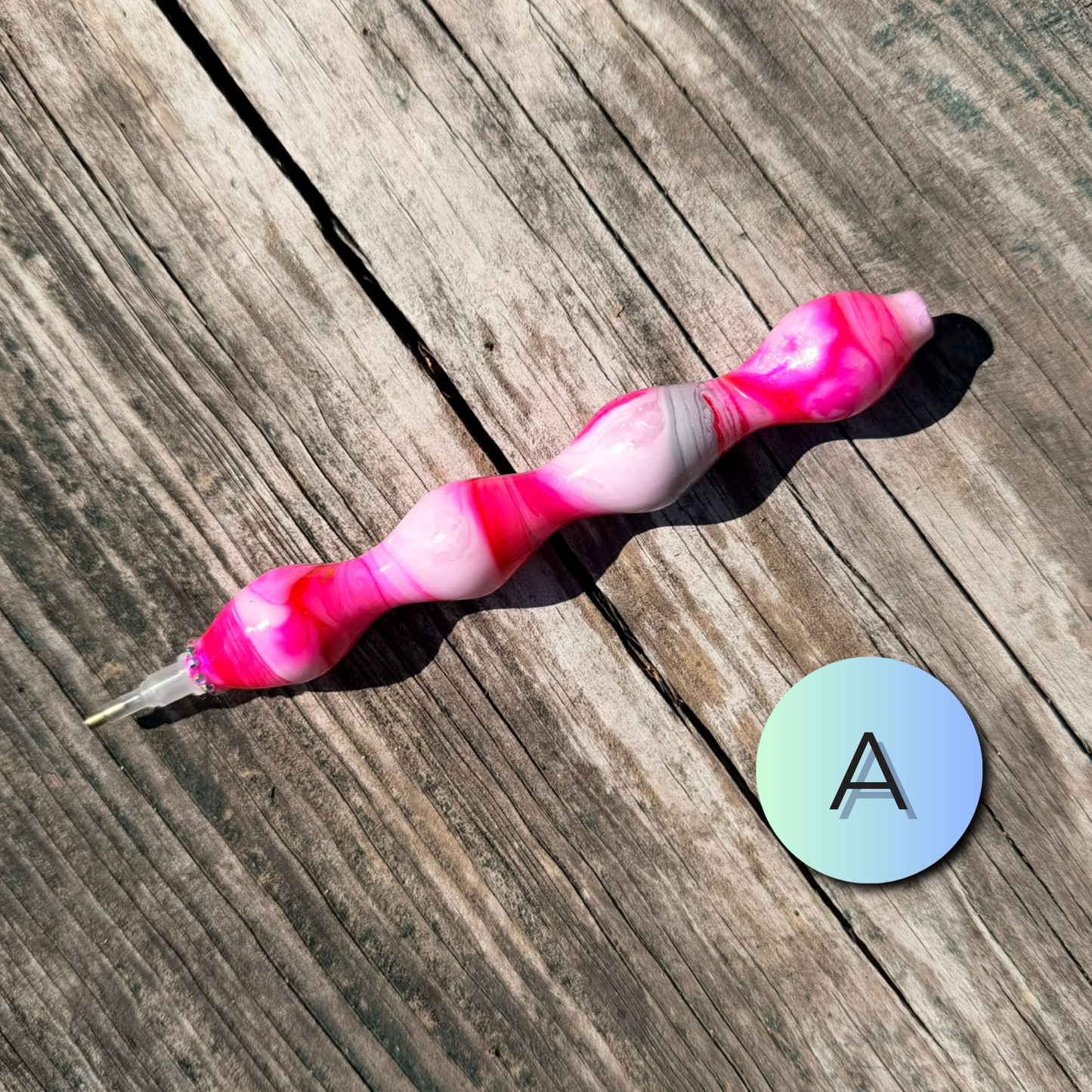 A marbled pink and white silicone nail art tool with a metal tip, placed diagonally on a weathered wooden surface next to a circular sticker with the letter 'A'.