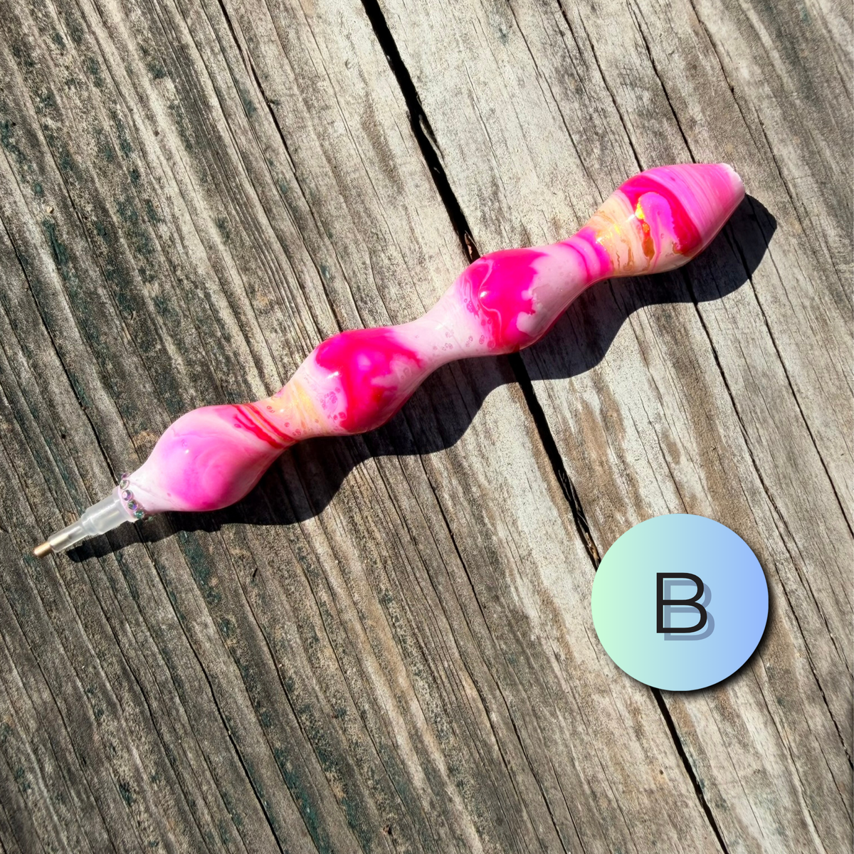 A marbled pink and white pen with a clear cap lies on a weathered wooden surface next to a circular blue-to-green gradient sticker featuring the letter 'B'.