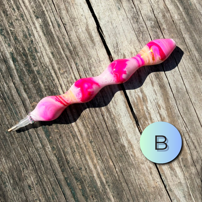 A marbled pink and white pen with a clear cap lies on a weathered wooden surface next to a circular blue-to-green gradient sticker featuring the letter 'B'.
