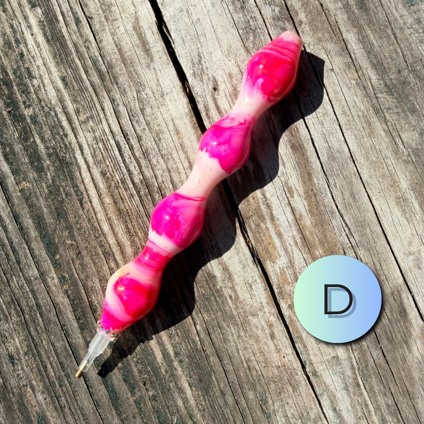 A pink and white marbled silicone personal massager with a clear tip, displayed diagonally on a weathered wooden surface next to a circular sticker with the letter 'D'.
