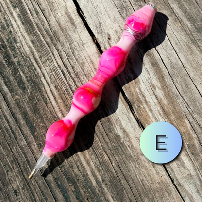 A pink and white marbled silicone personal massager with a clear suction tip, displayed diagonally on a weathered wooden surface next to a circular blue-green gradient sticker with the letter 'E'.