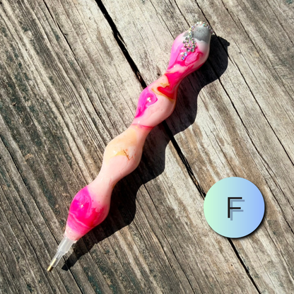 A pink and peach marbled silicone personal massager with a clear tip, displayed diagonally on a weathered wooden surface next to a circular blue gradient sticker with the letter 'F'.