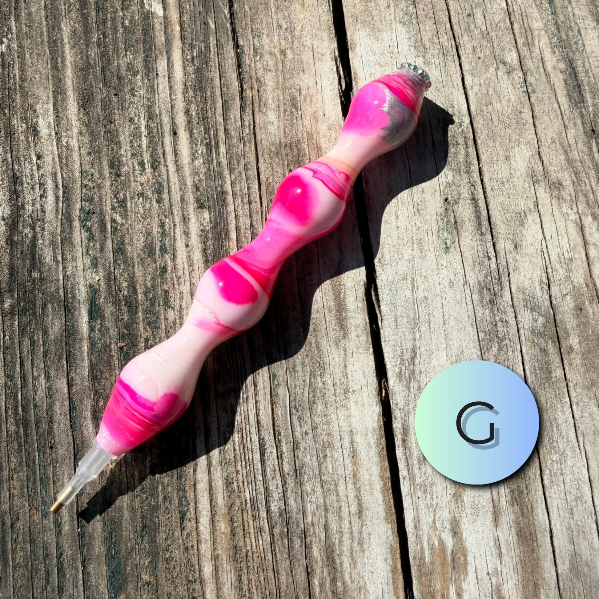 A pink and white marbled silicone pen-like tool with a clear tip, placed diagonally on a weathered wooden surface next to a round iridescent sticker with a black letter G.
