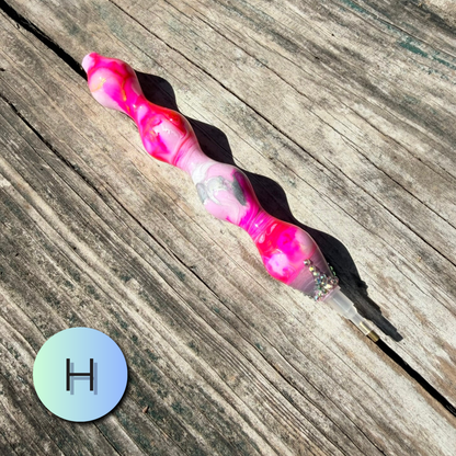 A pink and white marbled silicone pen with a clear end cap filled with small glitter, placed diagonally on a weathered wooden surface. A circular gradient icon with the letter 'H' is in the bottom-left corner.